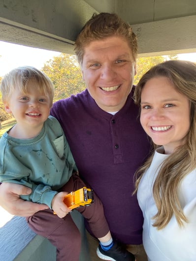 Man with sandy colored short hair in a purple shirt stands between a small blond child holding a yellow toy car and a blond woman with hair past her shoulders and wearing a white shirt.
