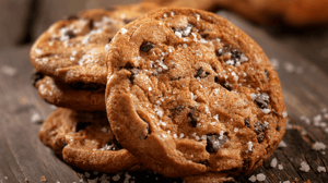 Close up shot of three chocolate chip cookies with sea salt sprinkled on top. Leaning on the stack is another of the same cookie. All cookies are on a wood counter and there are pieces of sea salt around the cookies.