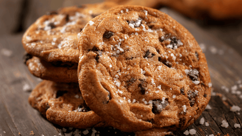 Close up shot of three chocolate chip cookies with sea salt sprinkled on top. Leaning on the stack is another of the same cookie. All cookies are on a wood counter and there are pieces of sea salt around the cookies.