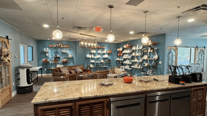Looking over an island that has tan granite countertop, brown lower cabinets and three dishwashers. On top of the counter are three blenders to the right. In the space in front of the island is leather seating and a wall full of shelves and smallwares.