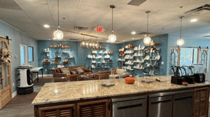 Looking over an island that has tan granite countertop, brown lower cabinets and three dishwashers. On top of the counter are three blenders to the right. In the space in front of the island is leather seating and a wall full of shelves and smallwares.