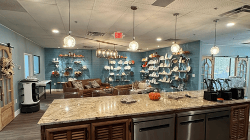 Looking over an island that has tan granite countertop, brown lower cabinets and three dishwashers. On top of the counter are three blenders to the right. In the space in front of the island is leather seating and a wall full of shelves and smallwares.