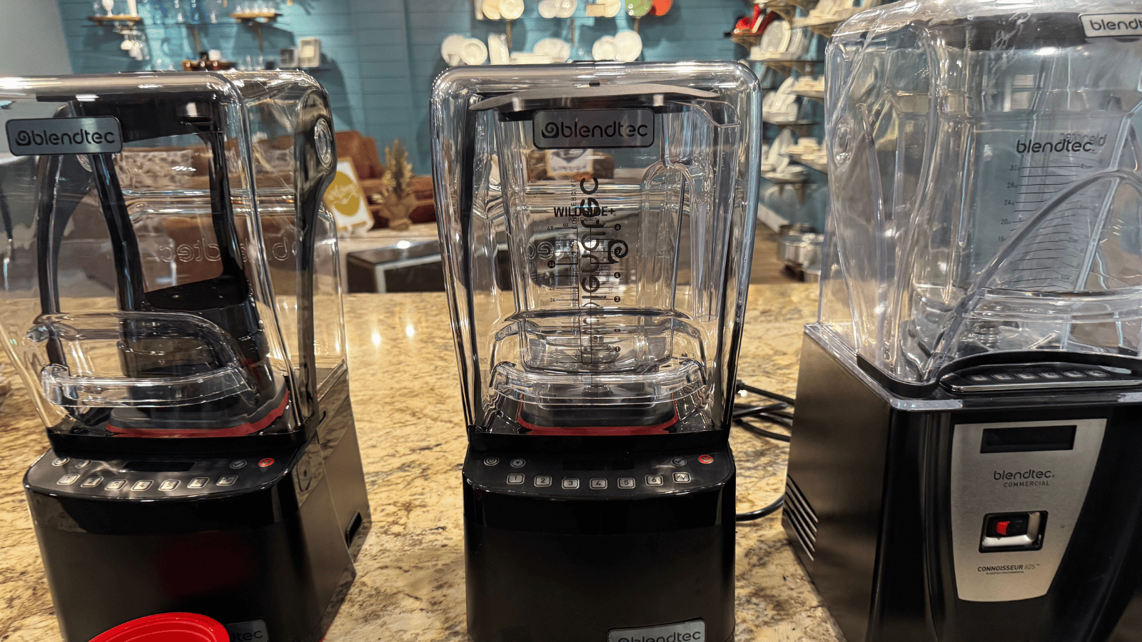Three Blendtec blender models sit on a tan granite countertop.  Blurred in the background is a wall with shelving full of smallwares including dishes.