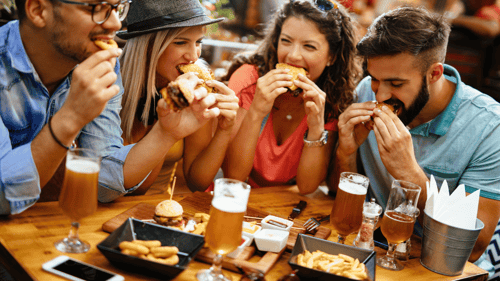 Two men and two women at a table eating slider burgers. Table has trays of burgers and fries, dipping sauces, bucket of napkins and glasses of beer.