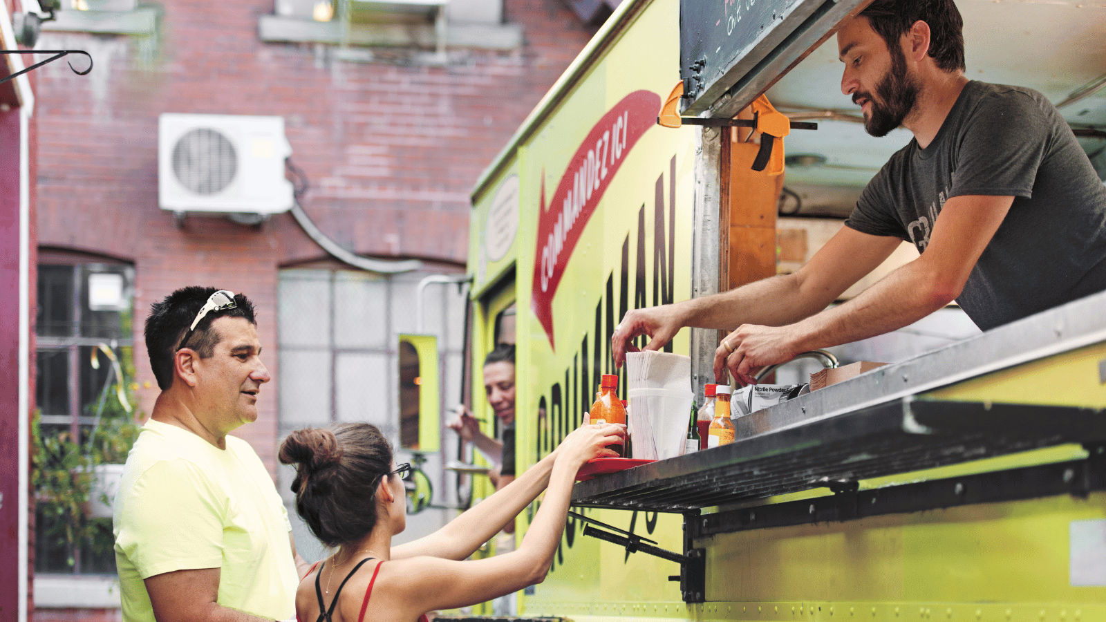 Man with dark hair and beard leans out of yellow food truck to hand woman in red top something. 