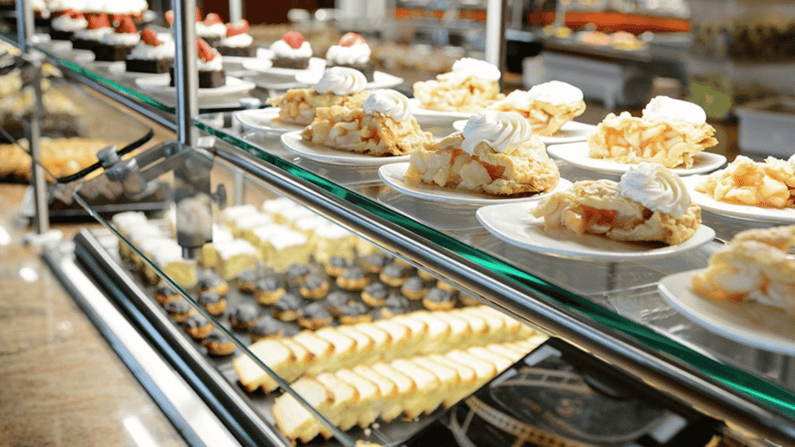 Glass food shield protecting plated desserts on a commercial serving line, with clear visibility and stainless supports in a high-volume foodservice environment.