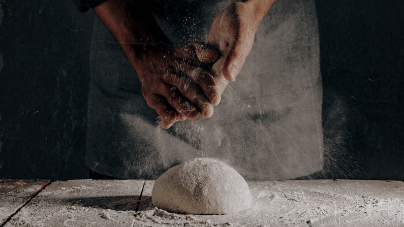 Close-up of hands shaping a dough ball on a floured surface, illustrating manual dough preparation and the consistency challenges automation helps solve.