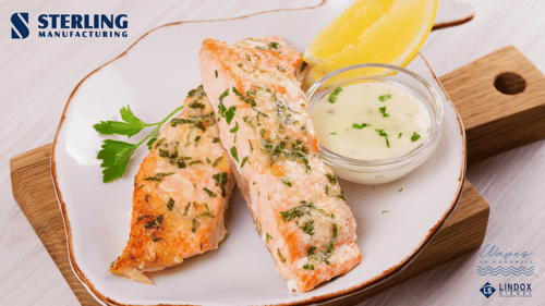 Two pieces of steamed salmon sit on a white plate with brown edging. On the plate is also a ramekin of white sauce, a slice of lemon, and a sprig of parsley. The plate sits on a brown cutting board. The Sterling logo is in the top left; the Lindox Siegel logo is in the bottom right.