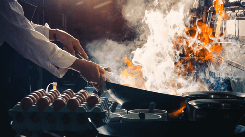 A large black wok is held above a gas stove in a restaurant kitchen. There are tall flames and a lot of smoke coming from inside the wok. A cardboard tray of two dozen uncooked eggs are on sitting on the edge of the stove.